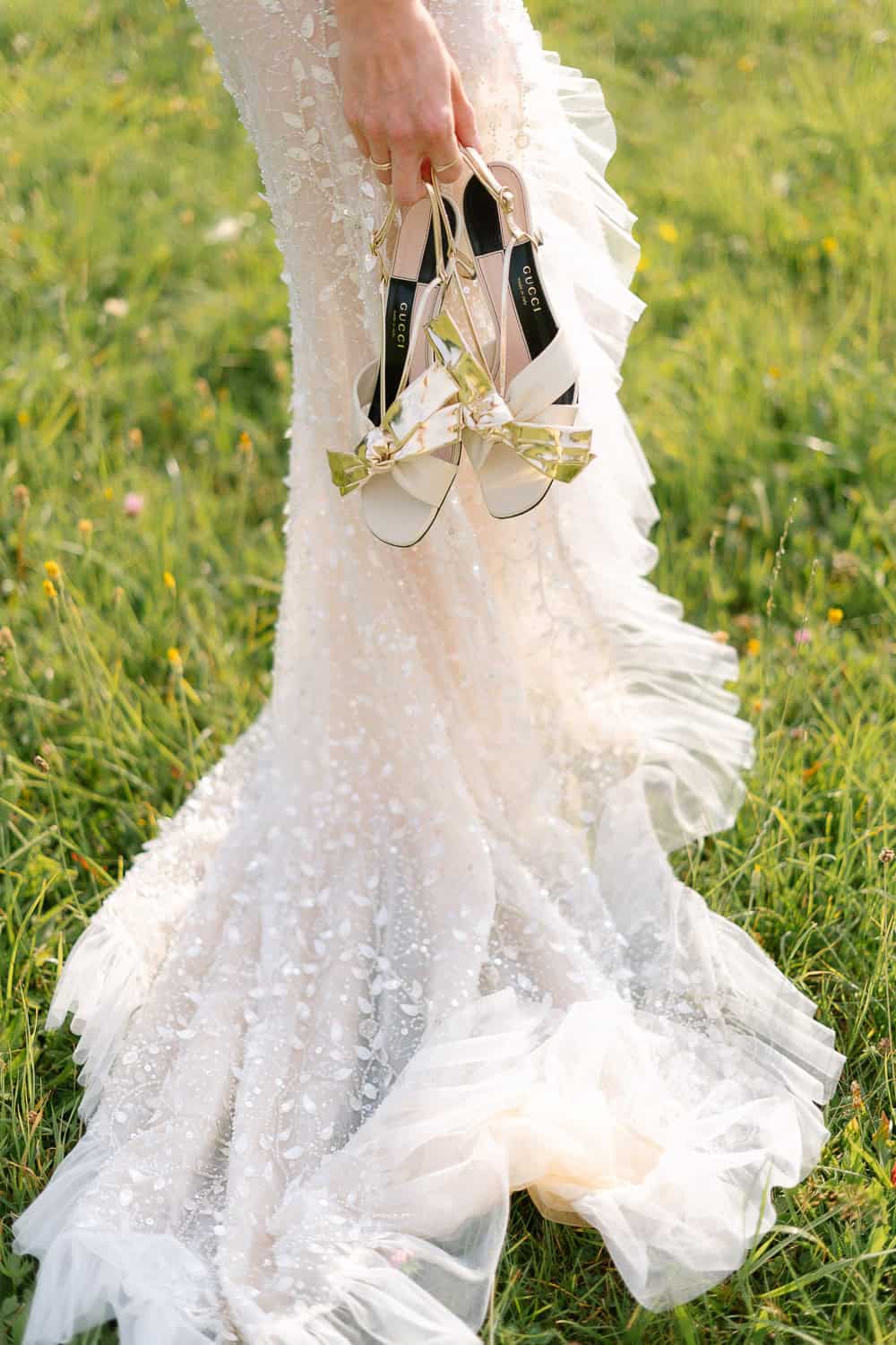 Braut im Spitzenkleid hält weiße Sandalen mit goldenen Schleifen.