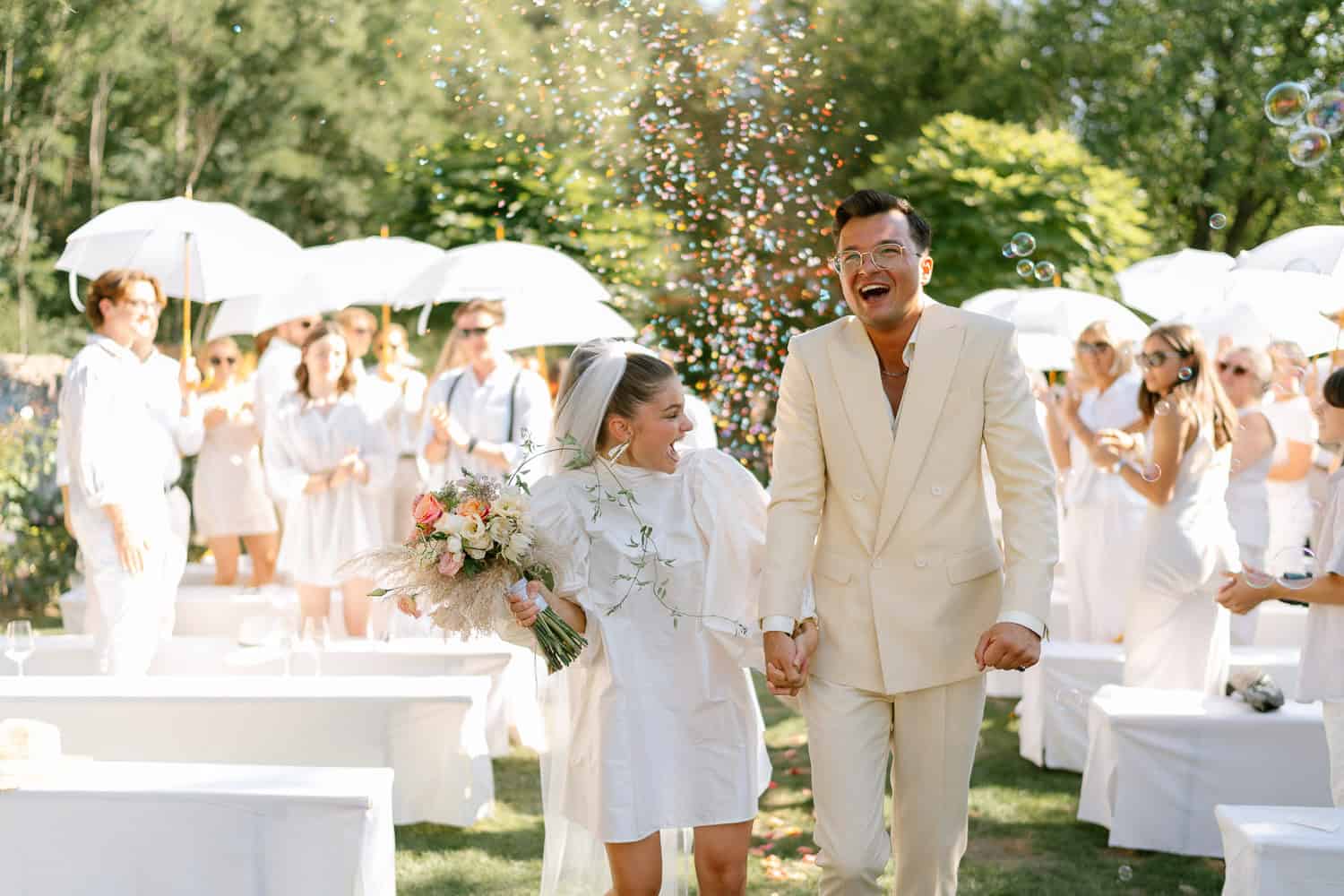 Hochzeitspaar lächelt im Freien, Gäste mit Regenschirmen und Konfetti. Hochzeit im Stadtflucht Bergmühle