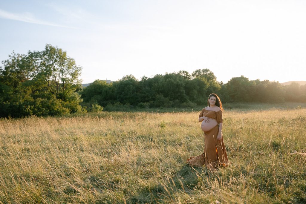 Schwangere Frau im braunen Kleid auf sonniger Wiese mit Bäumen.