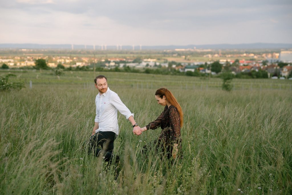 Paar hält sich im hohen Gras mit Stadt und Windrädern an den Händen.