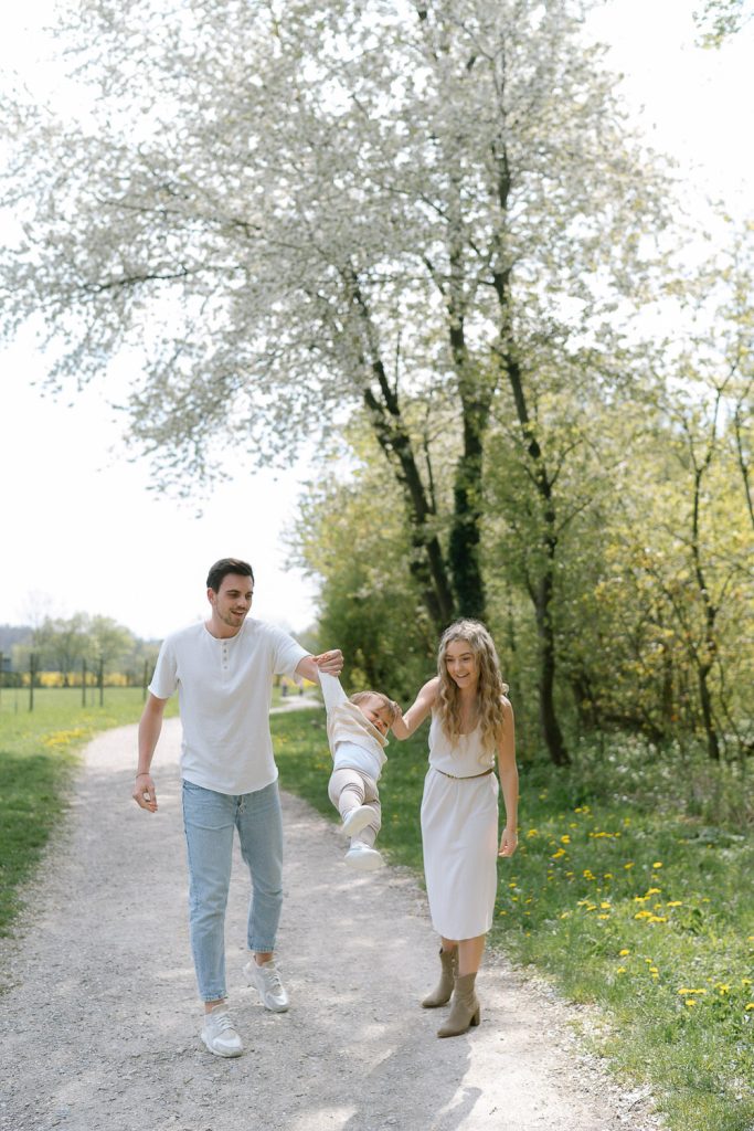 Familienfotoshooting: Eltern schaukeln Kind auf sonnigem Frühlingsweg.