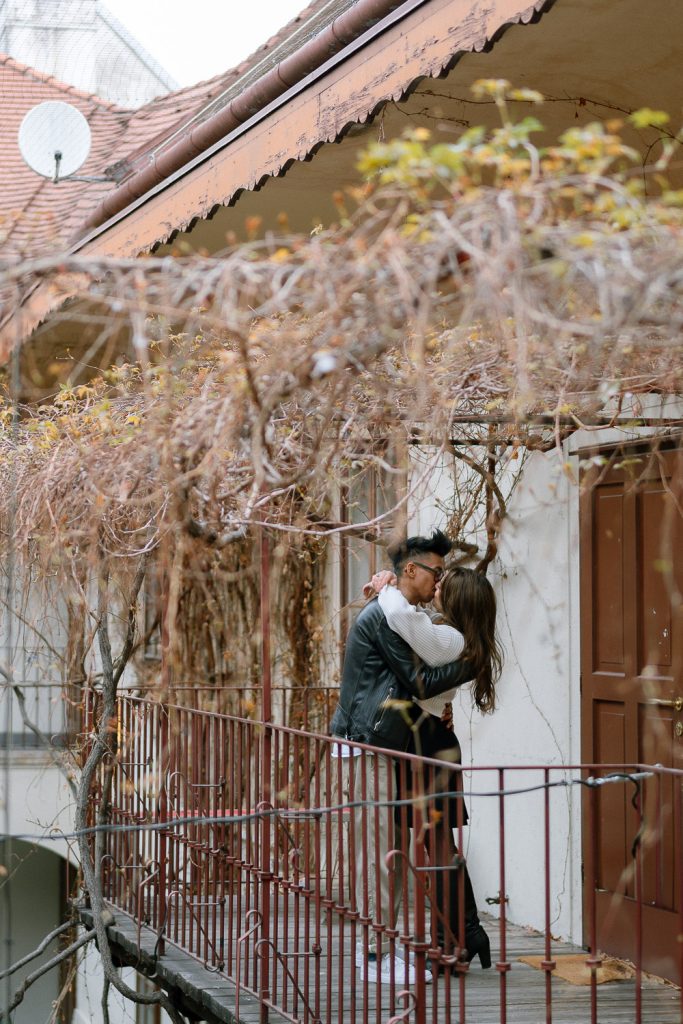 Ein Paar küsst sich auf einem Balkon mit roten Türen und blattlosen Ranken.