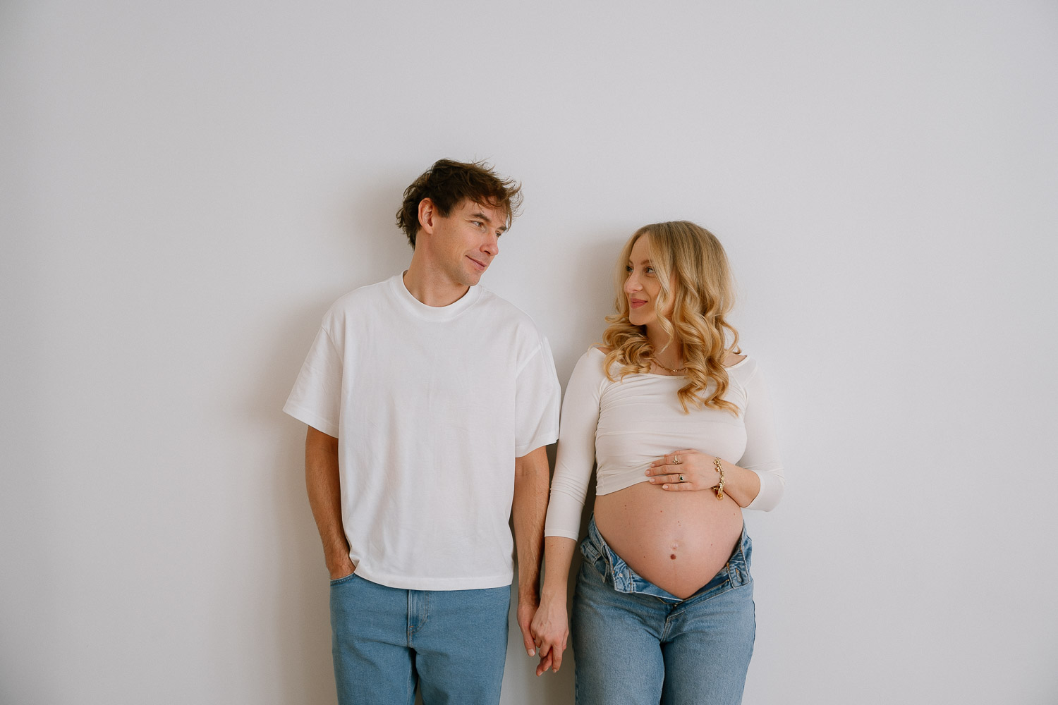 Lächelndes Paar in weißem Shirt und Jeans, Hand in Hand vor heller Wand – moderne Babybauchfotos in Wien