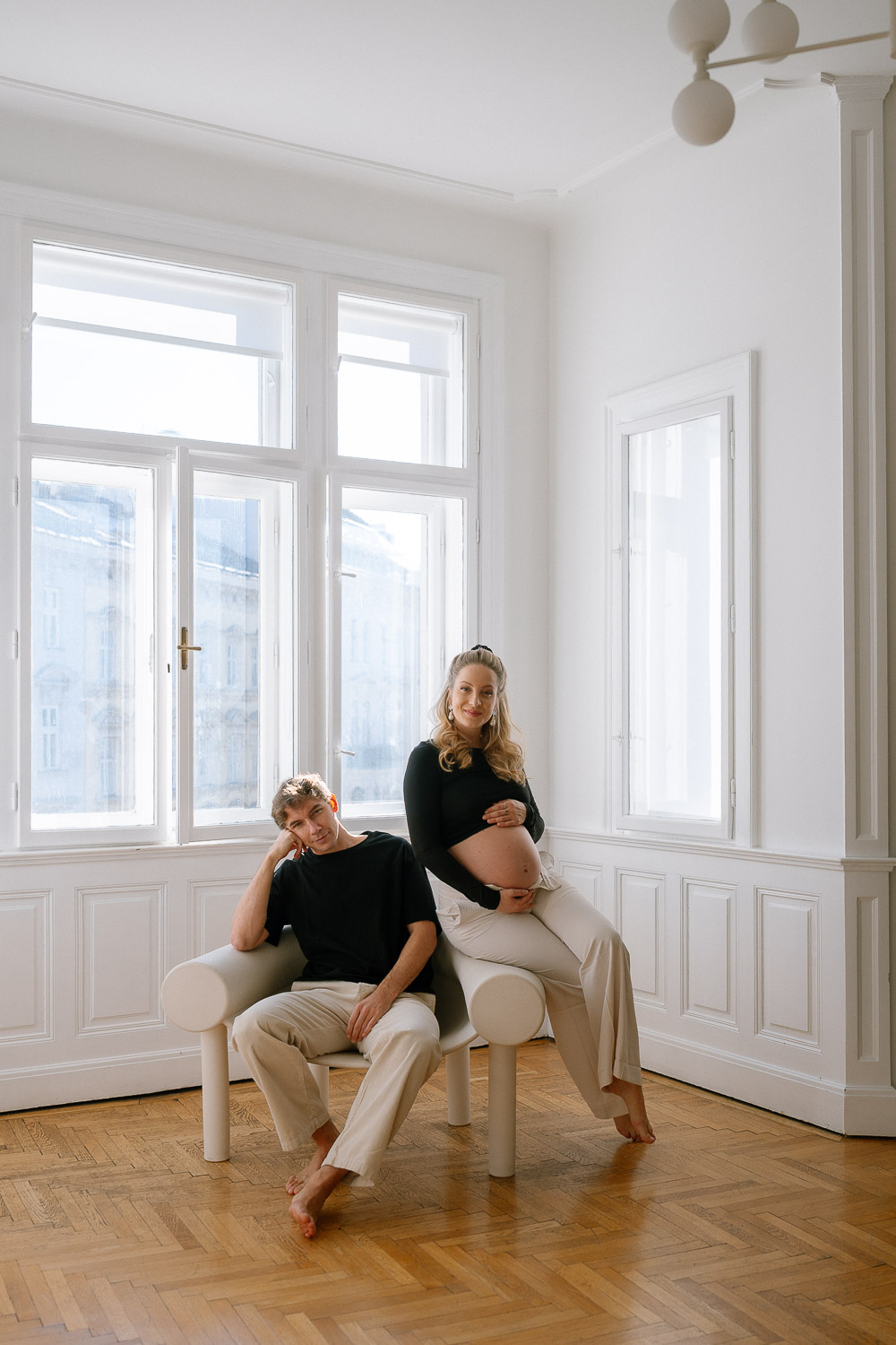 Paar sitzt entspannt auf einem Designer-Sofa vor großen Fenstern – moderne und stilvolle Schwangerschaftsfotografie in Wien