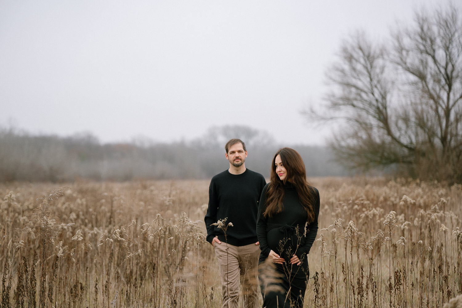 Ein Paar steht in einem nebligen Feld, beide in Schwarz gekleidet – die Schwangere im Vordergrund, der Partner im Hintergrund.