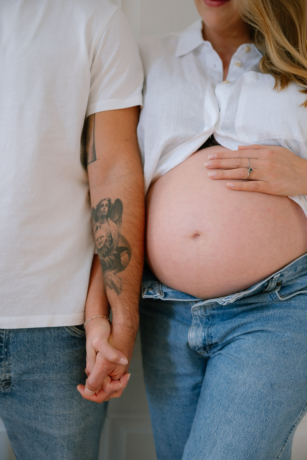 Detailaufnahme eines schwangeren Bauches – die Frau trägt ein weißes Hemd und Jeans, der Partner hält ihre Hand.