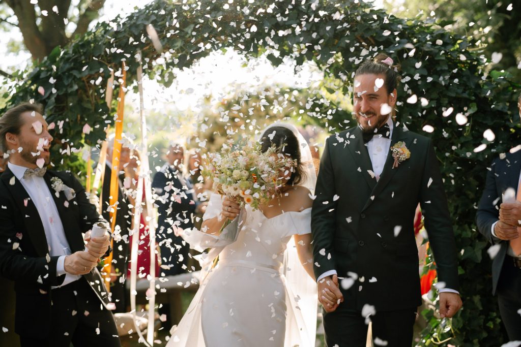 Braut und Bräutigam beim Auszug unter einem Regen aus weißem Blütenkonfetti nach der Trauung