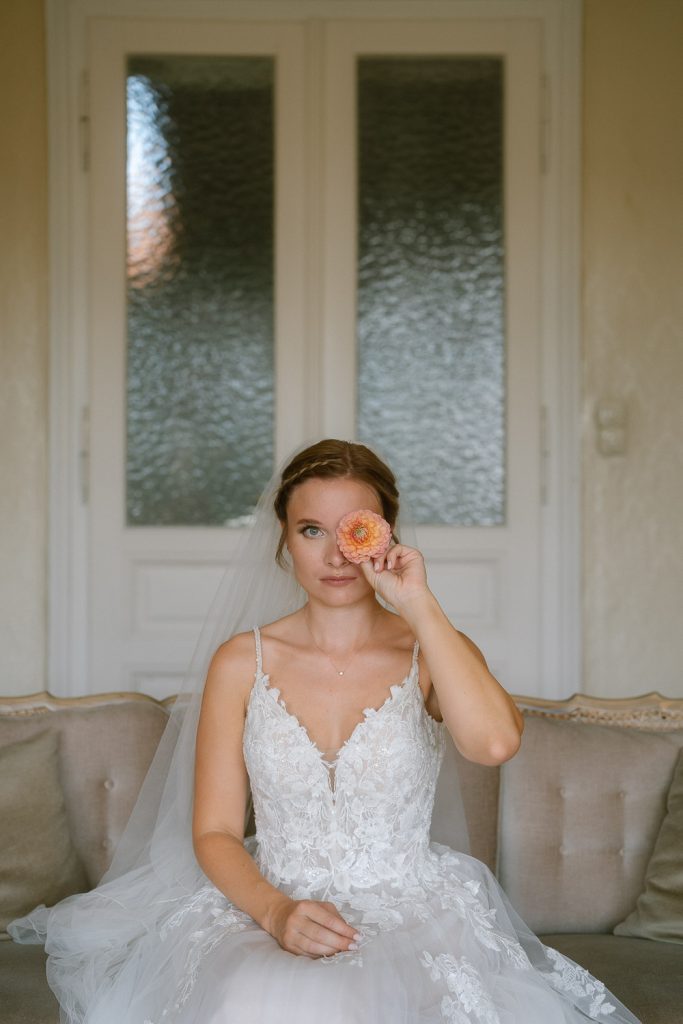 Braut sitzt in einem hellen Raum und hält eine Blume vor das Auge – verspieltes Porträt bei der Hochzeit am ROAN.