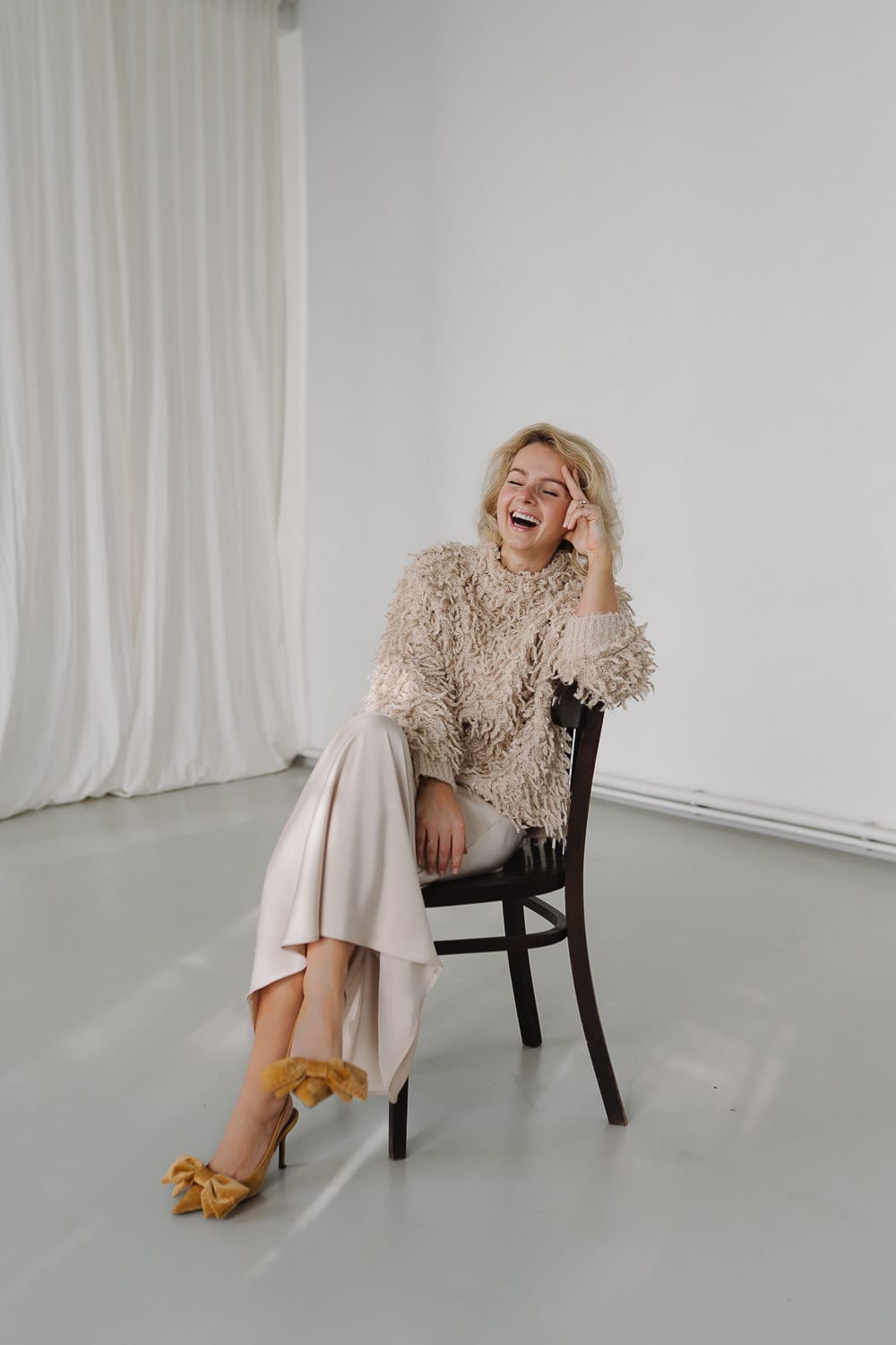 Blonde Frau lacht auf Stuhl, beige Outfit, helle minimalistische Fotografie.